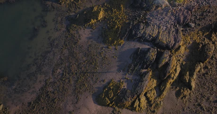 A lone person walking along the textured shoreline the person moves across a mix of dark sand and sunlit rock formations and a body of water is visible on the left side of the frame