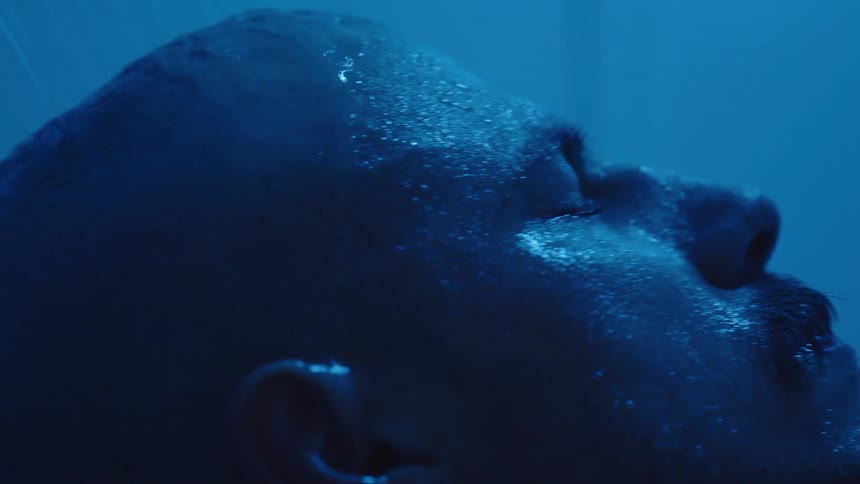 A close side view shows a mans washing is bald head and face in the shower with water droplets running down his skin against a dramatic deep blue background