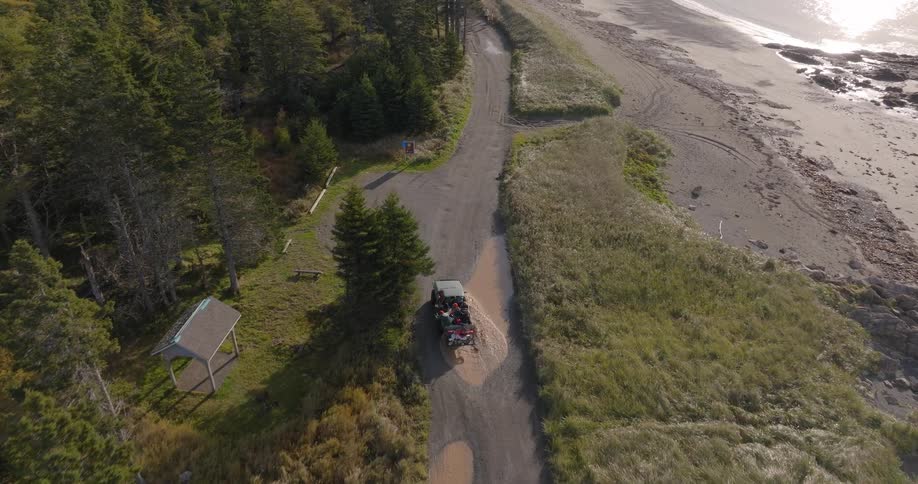 The video is an aerial shot of a dirt road running between a thick evergreen forest and a wide sandyshingle beach a small allterrain utility vehicle carrying passengers is driving slowly along the road away from the camera
