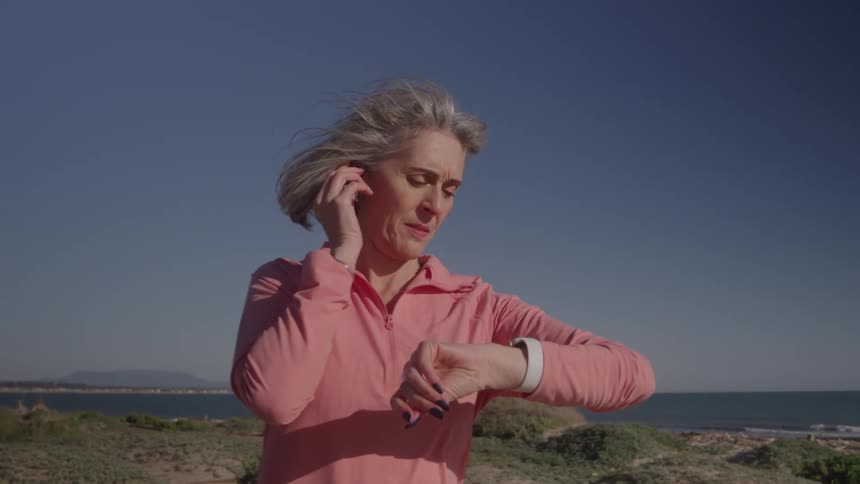 A middleaged woman with grey hair wearing a pink longsleeved shirt is shown in a close view looking at her smart watch and adjusting a wireless earbud while jogging outdoors on a sunny day near the ocean