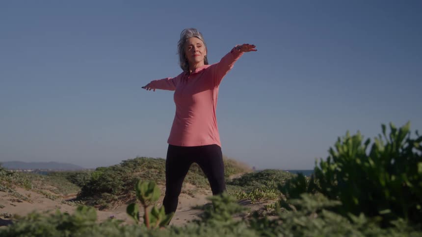 Grayhaired woman practices a balancing pose on the sand with the sea in the background