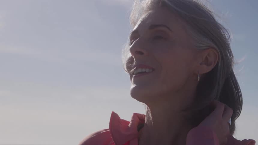 A close side view shows an older woman with grey hair smiling while looking up toward the sun raising her hand to touch her neck outdoors on a bright day
