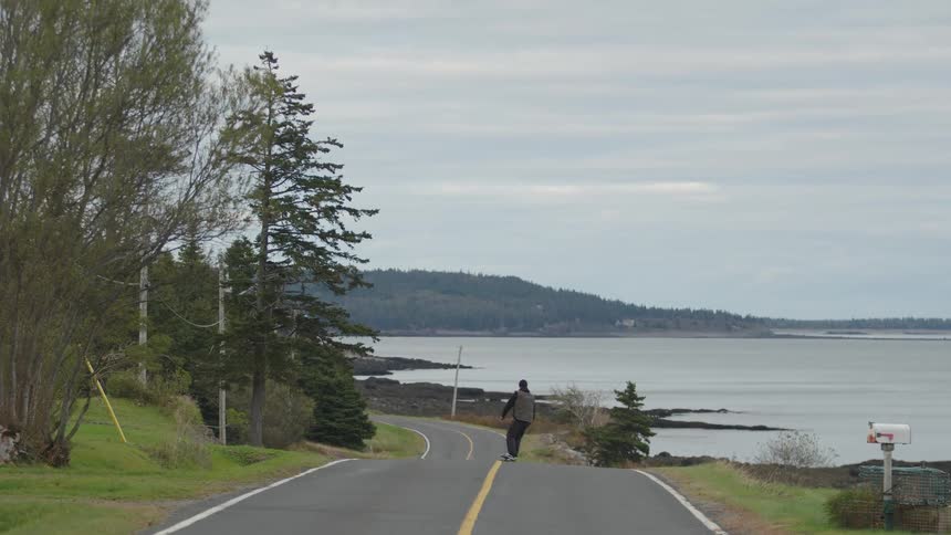 A man wearing a jacket rides a skateboard down a hill road at midday the lake shimmering in the background under the clear sky as he picks up speed along the descent