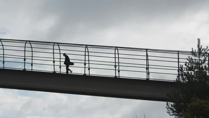 Person walks across a metal footbridge carrying a guitar case under a cloudy sky