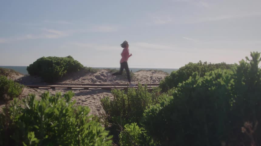 A woman runs across sand next to ocean passing by railroad tracks between low green plants under a bright sky