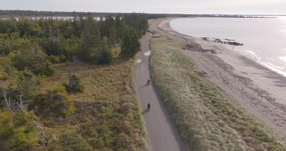 An aerial view follows two people on bicycles riding on a paved road that runs along a grassy shoreline next to a wooded area and the vast gray ocean under a pale sky