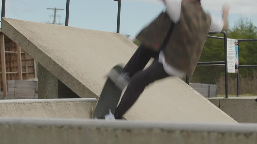 A low angle close view shows a skateboarder wearing a dark jacket and tan pants riding up a steep concrete ramp in a skate park on an overcast day