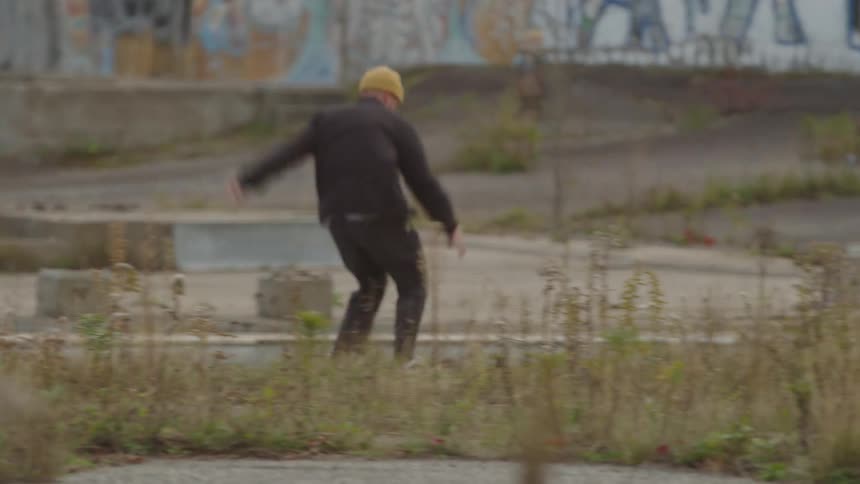 A man wearing a black jacket dark pants and a yellow beanie cap performs a small jump on a skateboard in an overgrown derelict urban area with concrete blocks and graffiti in the background