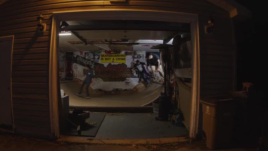 Three young adult male friends skateboarding together on a ramp in a garage at night