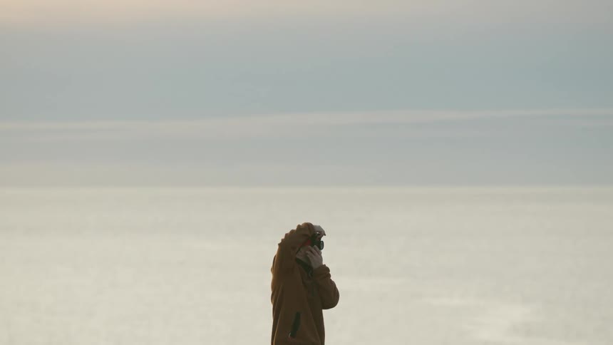 Individual standing by large body of water at sunset wearing brown jacket and holding optical device to face