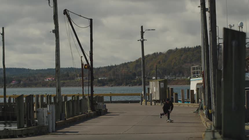 A man wearing dark clothing and a black hat skateboards down a gray wooden pier or dock surrounded by pylons with a forested hillside across the water under an overcast sky
