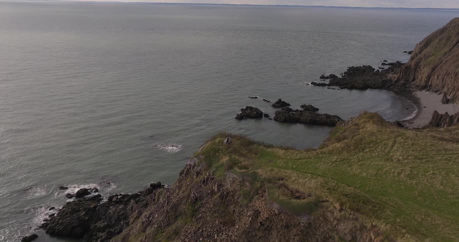 The video is an aerial shot of a rugged coastline where a grassy cliff meets a rocky shore and the ocean the gray sea is expansive under an overcast sky with waves crashing against the base of the cliffs