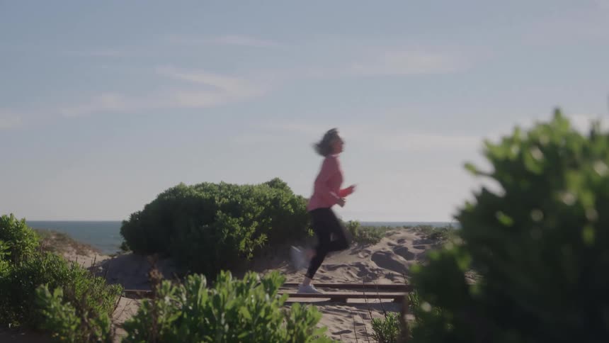 Woman running on a sandy trail surrounded by vegetation near the sea on a sunny day