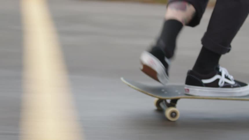A persons feet wearing black and white shoes are on a skateboard deck with wheels propelling the board forward on a gray asphalt surface with a yellow stripe