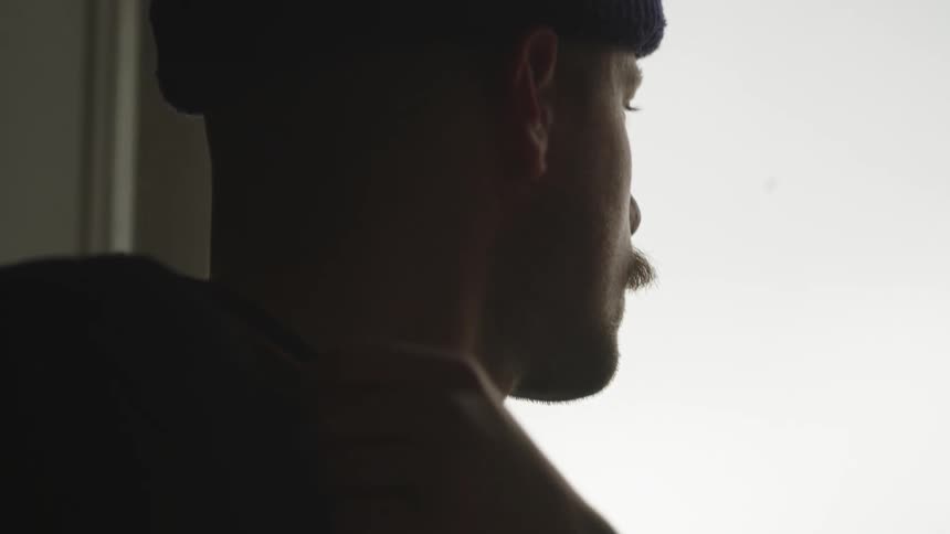 Man wearing a beanie keeps his eyes closed in silence near a bright window