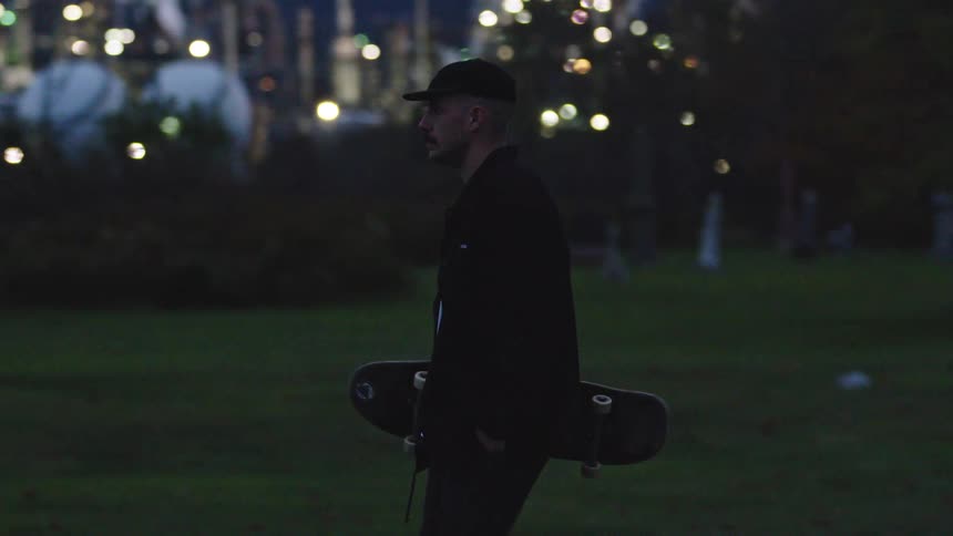 Man carries a skateboard through a park at dusk with city lights