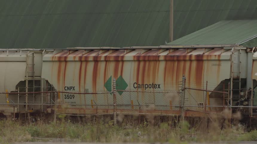 A medium view shows a rusted white train car with canpotex and cnpx 3509 stenciled on the side and a green diamond logo seen through a chain link fence in an industrial area with green storage buildings in the background