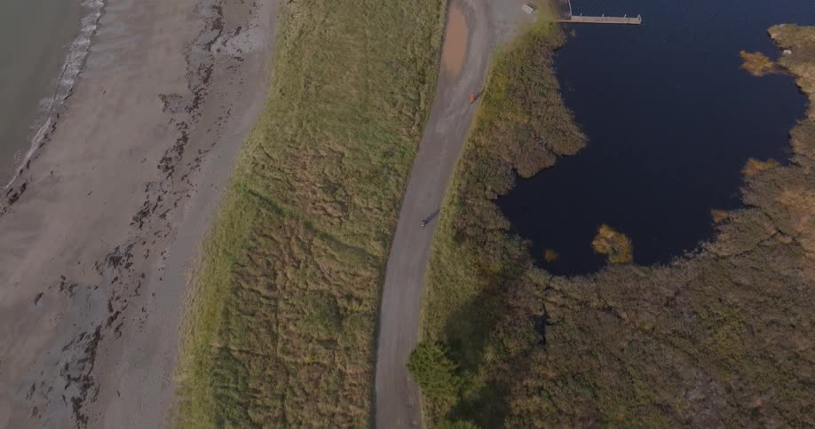 The video provides a high aerial view of a narrow unpaved path running between a wet sandy beachtidal flat and an adjacent dark body of water or marshland a single person on a bicycle can be seen riding along the path