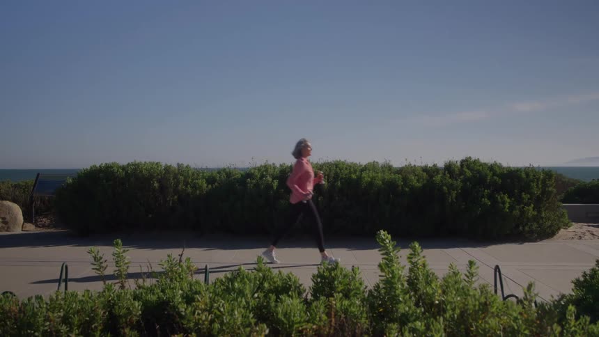 A woman in athletic wear jogs along a paved path next to a line of dense bushes and hedges with a wide view of the ocean and distant hills under a clear blue sky