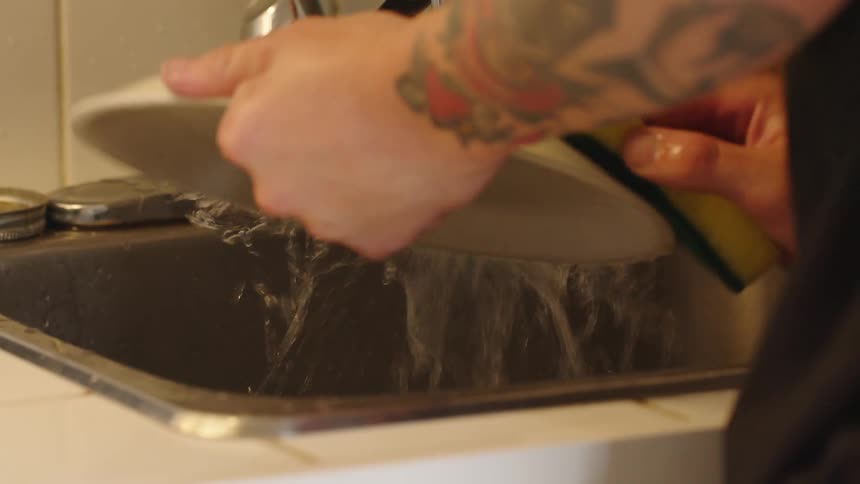 A close view shows the tattooed arm of a person rinsing a plate with a yellow and green sponge under running water in a stainless steel kitchen sink