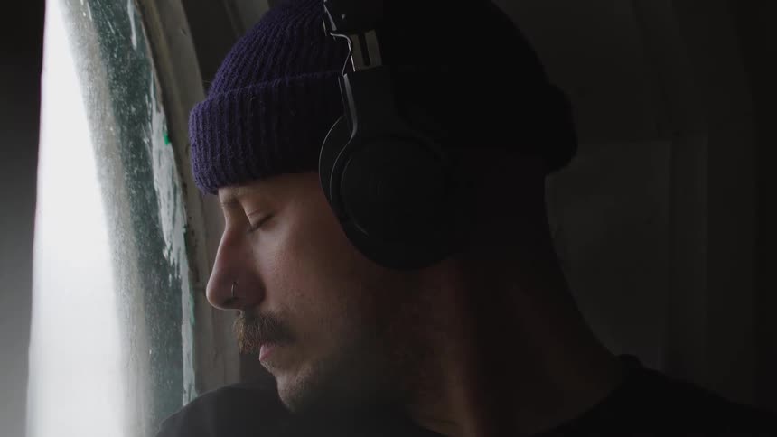 The video is a closeup profile shot of a man wearing a dark beanie and overear headphones he is standing in a dark room and looking out of a bright window with his face partially illuminated by the light