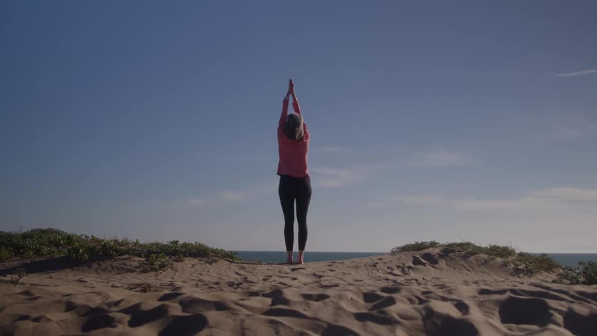Woman practices yoga on a sand dune gazing at the horizon under a clear blue sky
