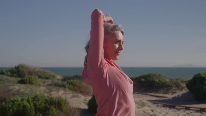 An active older woman in a pink top performs a tricep stretch outdoors with her arm overhead smiling faintly as she faces the ocean framed by sand dunes and blue sky