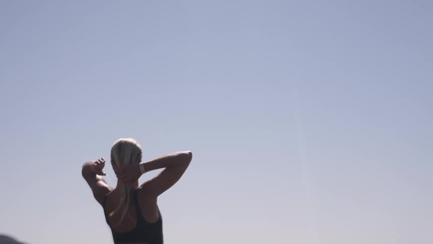 A view of a woman with blonde hair standing outdoors against a bright blue sky raising her hands to her head as if pushing her hair back