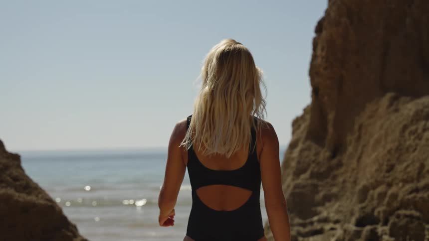 A medium shot from behind shows a blonde woman with long flowing hair wearing a black swimsuit with an open back standing between two sunny rock faces and looking out at the sparkling ocean