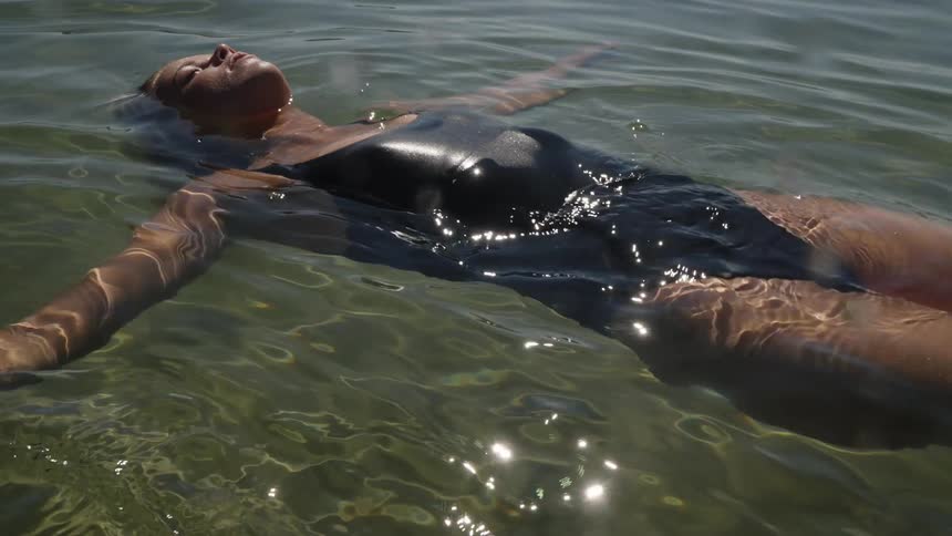 A person wearing a dark swimsuit floats face up on the surface of the water the arms are extended out to the sides the water is clear and shimmering with reflected sunlight and the person appears relaxed on a sunny day