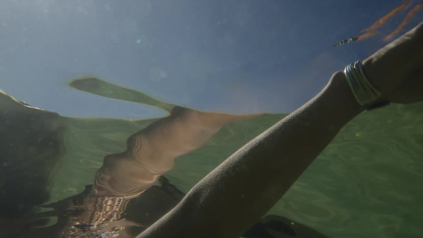 An underwater view in murky green water shows a woman wearing a dark swimsuit and silver bracelets swimming toward the surface under a blue sky reflected and distorted above the water line