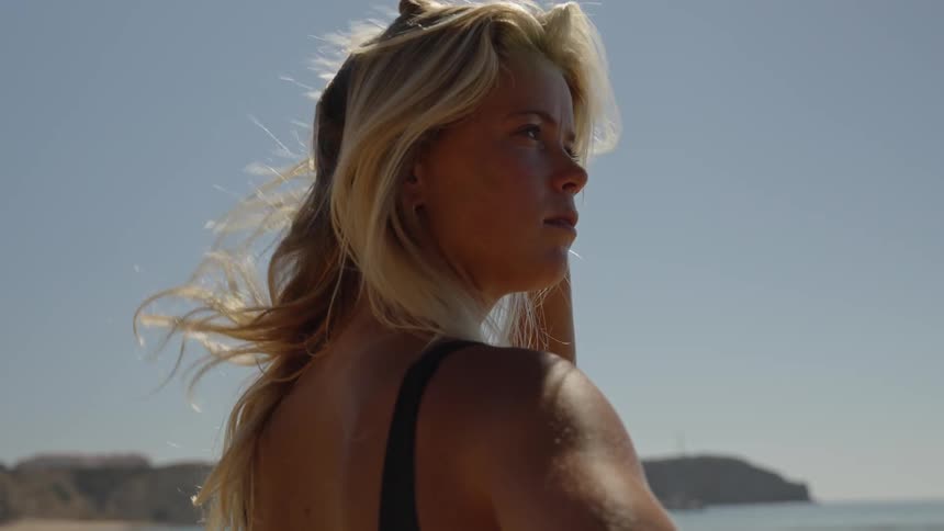 A woman stands in profile on a beach under a bright sun looking out over the water and slowly turning her head toward the camera