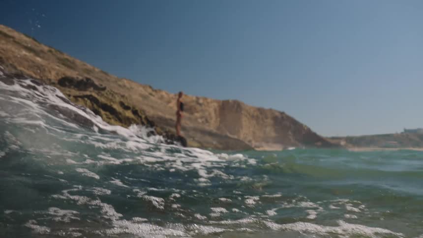 Woman standing on seaside rocks with waves crashing and cliffs in the background