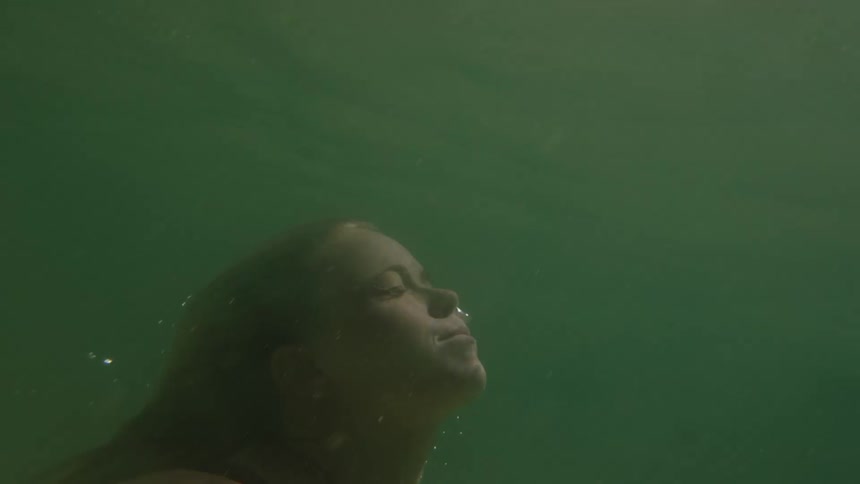 A person moves upward through green water their head and shoulders appear closer to the camera the persons face looks toward the surface the person breaks the water surface sunlight and water splashes are visible