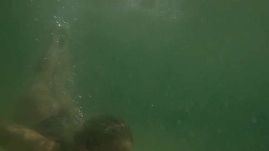 An underwater view in murky green water shows a woman wearing a dark swimsuit submerging and then slowly looking up toward the surface as bubbles float past her face