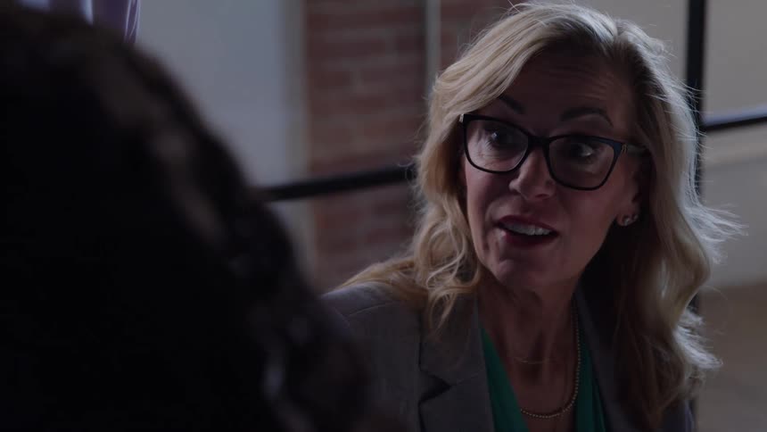 The video features a medium closeup shot of a blonde woman wearing glasses and a blazer speaking with an intense or animated expression to an unseen person across from her her face shows clear emotion as she talks in what appears to be an indoo