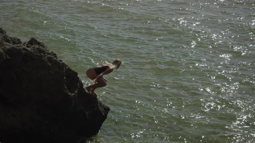 Blonde woman dives off a stone promontory into the ocean on a summer day