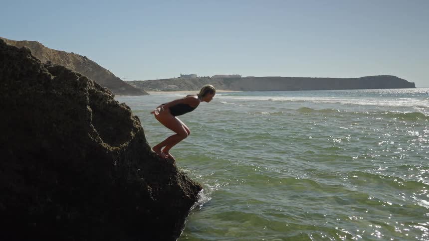 A young woman dives from a rocky cliff into the ocean swimming on a sunny day