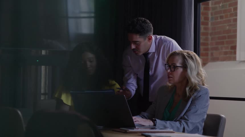 Man points at laptop screen during discussion with colleagues in office setting with large windows and brick wall
