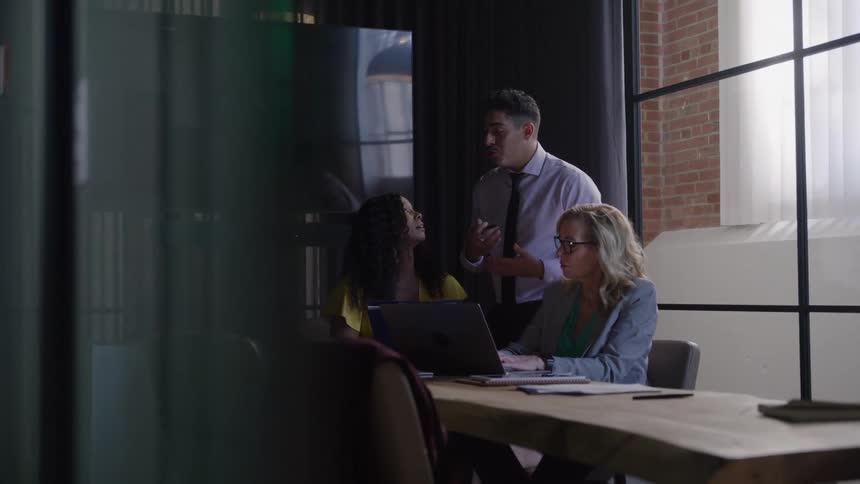 Three business people a man in a dress shirt and tie and two women in business casual attire are seen in a slightly obscured view having a collaborative meeting around a laptop at a wooden conference table in a modern office with large industrial window