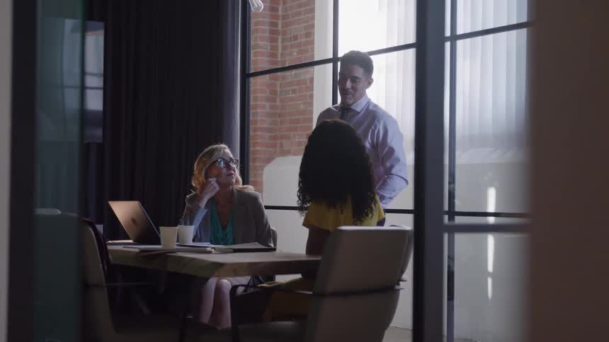 Three professionals a man in a tie and two women in business attire are seen through a glass office partition as they meet around a laptop at a table in a modern loft office with large windows and a brick accent wall