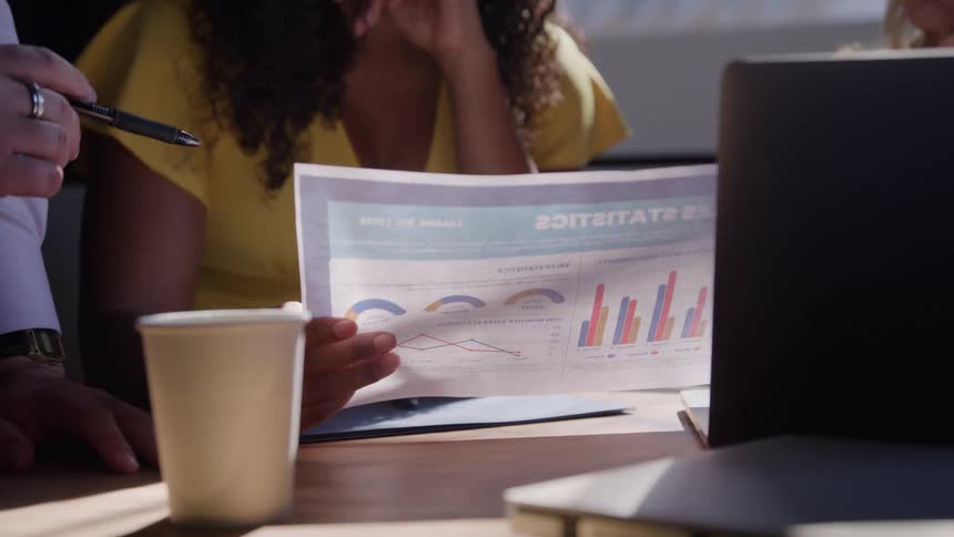 People sit around a table a paper cup rests on the table a persons hand holds a document showing graphs another persons hand points at the document a laptop sits on the right and people wear business attire