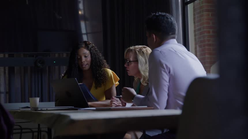 Colleagues discussing work over a laptop at a table in a dim office