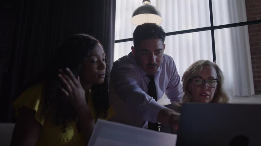 Three people sit and stand around a laptop a man leans over and points at the computer screen two women look at the screen and papers a window with curtains is behind the people and an overhead lamp hangs in the frame