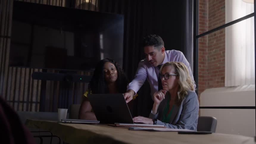 The video shows an indoor scene with a team of three people working around a laptop at a large table in an office setting the lighting is subdued and the backdrop features large windows and dark modern paneling