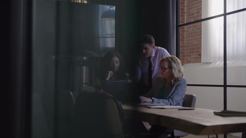 Three people sit and stand around a wooden conference table in an office room with brick walls and large windows looking at a laptop screen during a work interaction