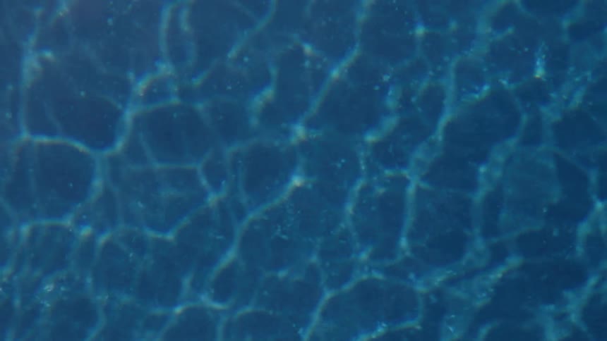 An underwater view shows the abstract pattern of bright sunlight shimmering and refracting on the dark blue surface of a swimming pool or the ocean floor