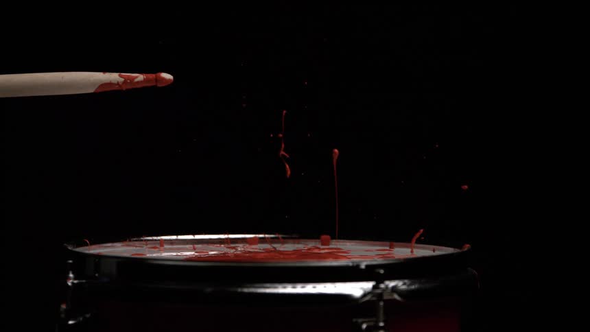 A stick strikes a band sending red paint splashing upward in a dark room