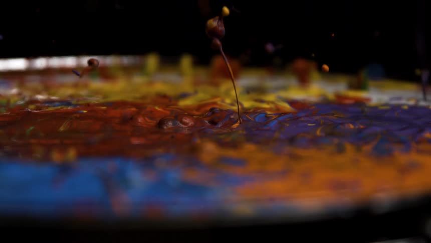 A close abstract view shows thick streams of dark blue orange and purple liquid resting onto a drum head with splashes of liquid paint flying upwards as it is impacted creating a contrasting pattern of color against a dark background