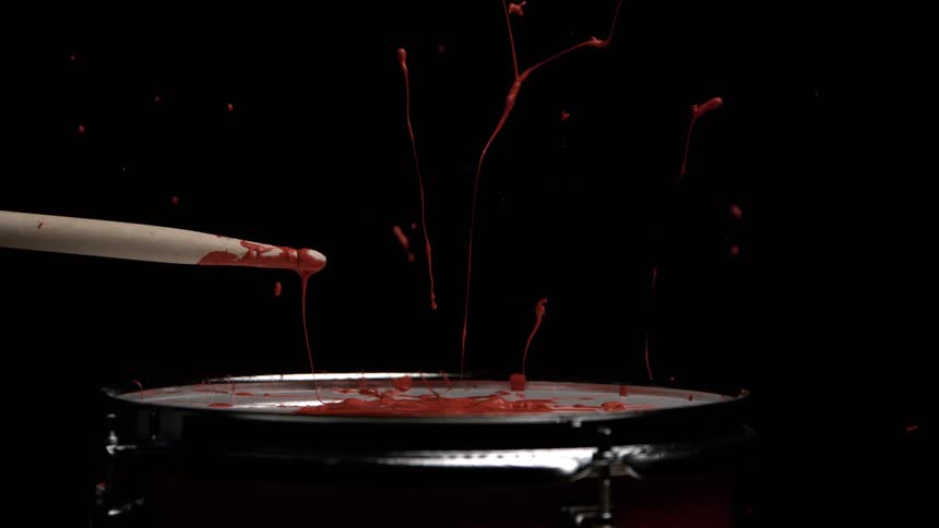 A wooden drumstick hits a drum head covered in thick orange liquid causing the substance to splash high in long thin strands against a solid black background
